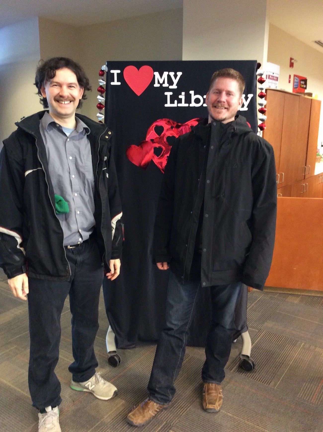 Two members of the University's IT department stand in front of the I love my Library backdrop wearing outerwear and smiling for the camera